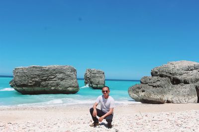 Full length of man on rocks at beach against clear blue sky