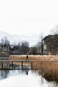 Side view of unrecognizable person walking on wooden boardwalk over calm lake with grassy shore in small settlement located in mountainous terrain in autumn day