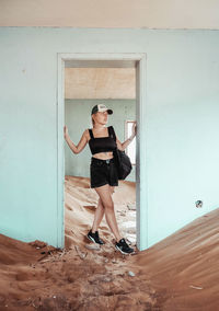 Full length of young woman standing against wall in a sandy house