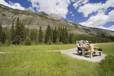 Family sitting on picnic table by mountain against cloudy sky