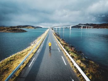 Rear view of man walking on road over sea against cloudy sky