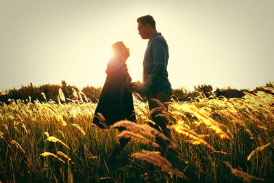 Side view of couple standing amidst grassy field against sky during sunset