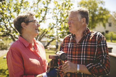 Happy senior couple holding hammers while looking at each other during sunny day in yard