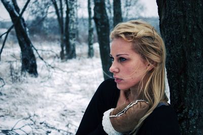 Thoughtful beautiful woman standing by tree trunk in winter