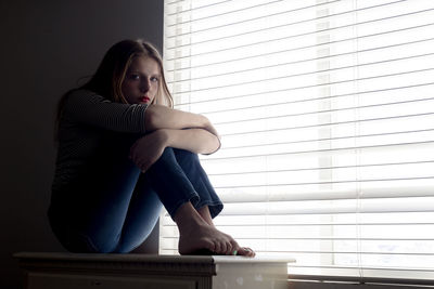 Full length portrait of woman sitting by window blinds on table