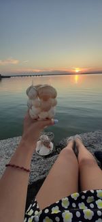 Low section of woman holding drinking glass against sky during sunset