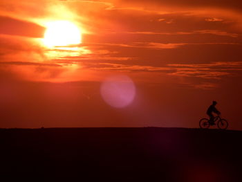 Silhouette man riding bicycle on field against orange sky