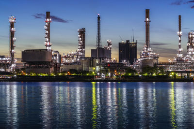 Illuminated factory by river against sky