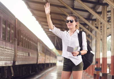 Young woman waving hand while standing at railroad station platform