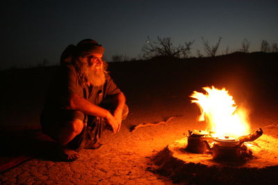 Mature man crouching at campfire on field during dusk