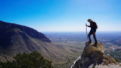 Young male hiker standing on rock against clear blue sky