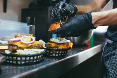 Side view of crop anonymous cook in black gloves arranging stick into freshly made hamburger while preparing order at kitchen counter