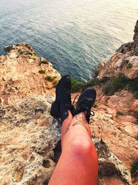 Low section of woman wearing shoes while sitting on cliff against sea