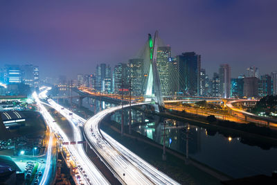 Light trails on road by illuminated buildings against sky at night