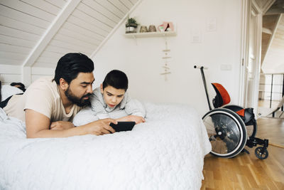 Father and disabled son watching movie over mobile phone while lying on bed at home