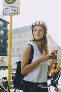 Young woman wearing bicycle looking away while holding smart phone in city