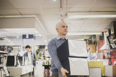 Mature salesman with boxes looking away while walking in electronics store