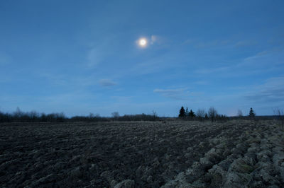 Plowed field and hills with green grass on horizon, blue sky and rising moon, foggy evening.