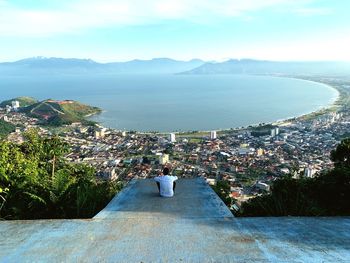 Rear view of man looking at cityscape by sea against sky