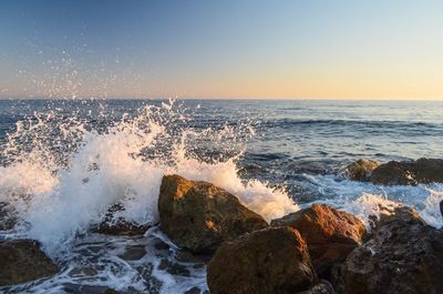 Waves splashing on rocks at shore against clear sky