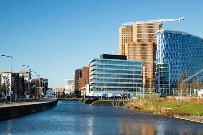 Modern buildings by river against clear blue sky