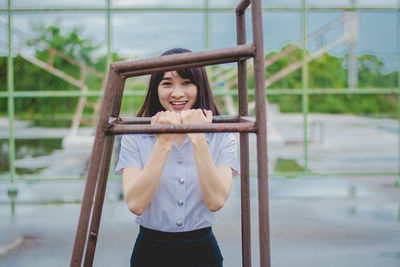Portrait of smiling young woman standing by metal at playground during rainy season