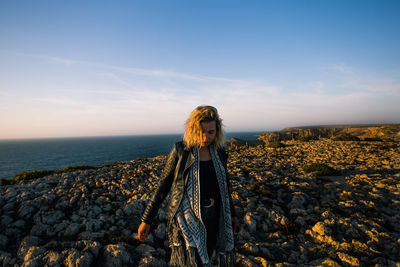 Full length of young woman standing on rock by sea against sky during sunset