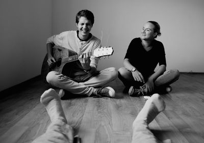 Young man playing guitar by friends on hardwood floor at home