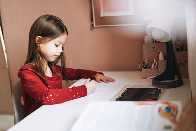 Pretty school girl with long hair in red dress draws with pencil at table in children's room at home