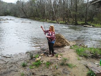 Full length of woman with fishing rod standing by river in forest