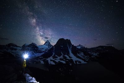 Hiker with illuminated headlight standing amidst mountains against sky at night