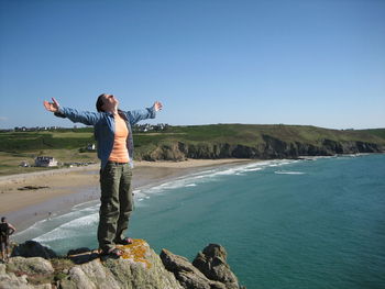 Young woman standing on cliff by sea against clear blue sky