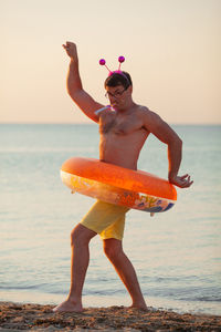 Full length of shirtless man with inflatable ring standing at beach during sunset