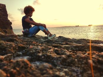 Woman sitting on rock at beach against sky during sunset
