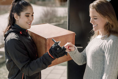 Female customer signing on smart phone while receiving package from delivery woman