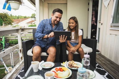 Father sharing tablet pc with daughter sitting on chair at porch