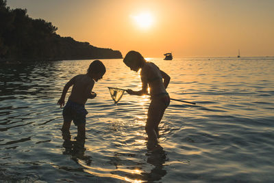 Silhouette boy and girl standing in sea during sunset