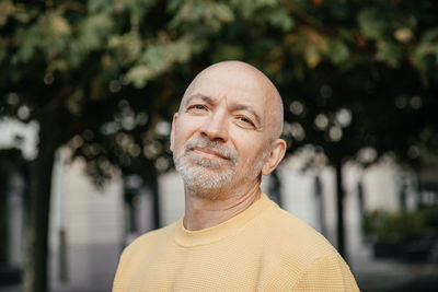 Content senior man in yellow sweater smiling gently in a tree-lined urban setting