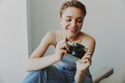 Portrait of young woman holding camera against wall