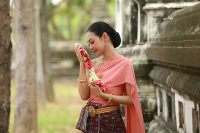 Young woman in traditional clothing holding flowers while standing against wall