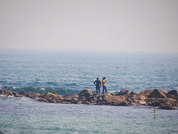 People standing on rocks at sea against clear sky