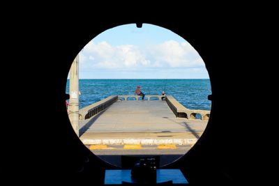 Man sitting at pier by sea against sky seen through circle window