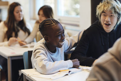 Happy teenage boy sitting by cheerful male friend at desk in classroom