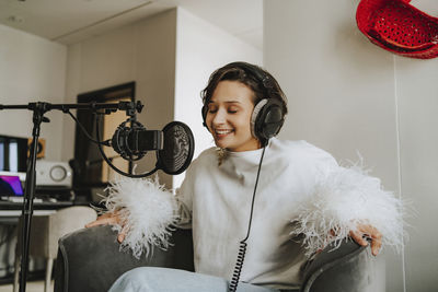 Smiling musician with eyes closed recording podcast through microphone on armchair in studio