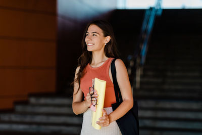 A smiling student girl stands on the steps and holds folders, glasses and a phone in her hands