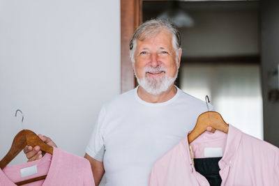 Glad mature male with gray hair holding hangers with shirts and looking at camera while preparing for festive event in apartment
