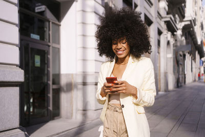 Smiling woman with curly hair using smart phone walking on sidewalk