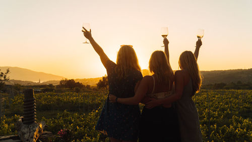 Rear view of women standing on landscape against sky during sunset