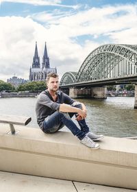 Portrait of mid adult man sitting on railing against hohenzollern bridge