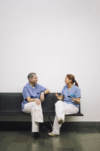 Smiling female trainee discussing with senior male doctor sitting on chair against white wall in hospital